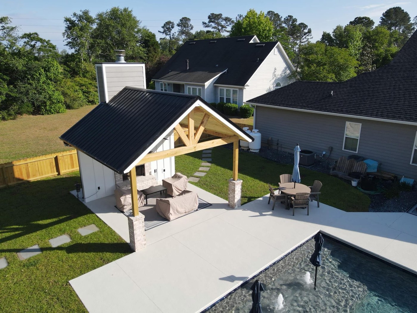 Custom poolside pavilion with a timber-truss gable, black metal roof, and outdoor fireplace, built by ProOps Constructs